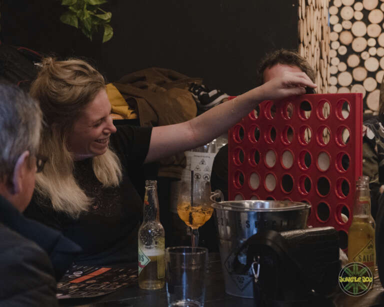 A group of friends enjoying a game of Connect 4 on a wooden table, with a smiling woman participating and drinks nearby.