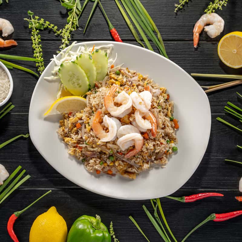 Stir-fried rice with eggs, onion and mixed vegetables on a white plate, on a black table decorate with vegetables around it.