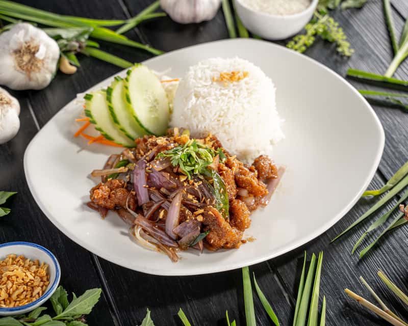Stir-fried crispy chicken in oyster sauce garlic and spring onion with a portion of rice on a white plate, on a black table decorate with vegetables around it.
