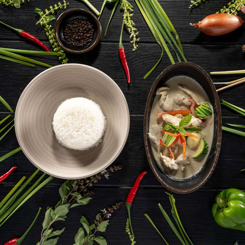 Thai green curry paste in coconut milk with zucchini, peppers and sweet basil with a portion of rice on a black table decorate with vegetables around it.