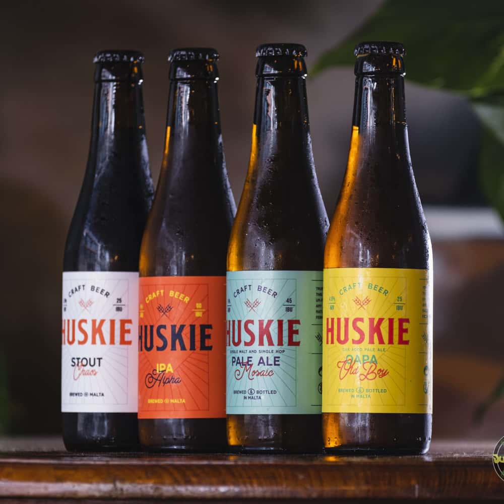 Four craft beer bottles featuring the brands 'Huskie Stout,' 'Huskie IPA Alpha,' 'Huskie PALE ALE Mosaic,' and 'Huskie OAPA Old Boy' arranged on a wooden table, with a blurred background.