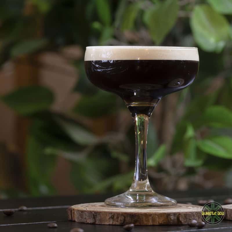 An espresso martini cocktail sits on a black wooden table with a tree in the background. The drink is a dark brown color and has a frothy cream top. It is served in a clear martini glass with a thin stem and wide bowl. The cocktail is garnished with three coffee beans resting on top of the cream. The lighting is dim, creating a moody atmosphere.