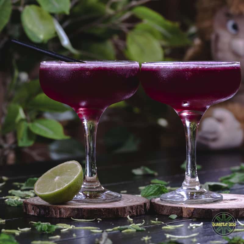 Two cosmopolitan cocktails are pictured on a black table with a blurry bokeh background. The drink is served in a manhattan glass and has a bright purple color. The liquid is clear and slightly cloudy. A thin long stirrer stick is placed inside the glass. The lighting is soft and creates a warm glow around the glass.