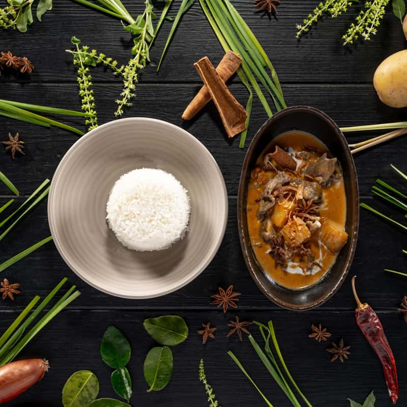 Massaman curry with coconut milk, potatoes, onion and roasted peanuts with a portion of rice on a black table decorate with vegetables around it.