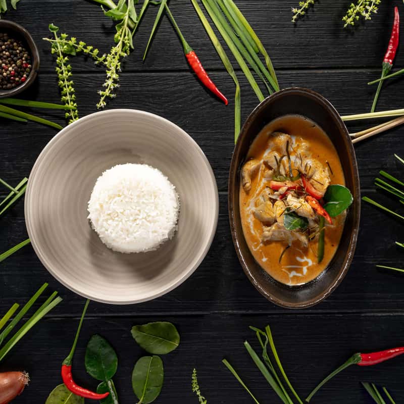 A rich curry of coconut milk with kafir lime leaves with a portion of rice on a black table decorate with vegetables around it.