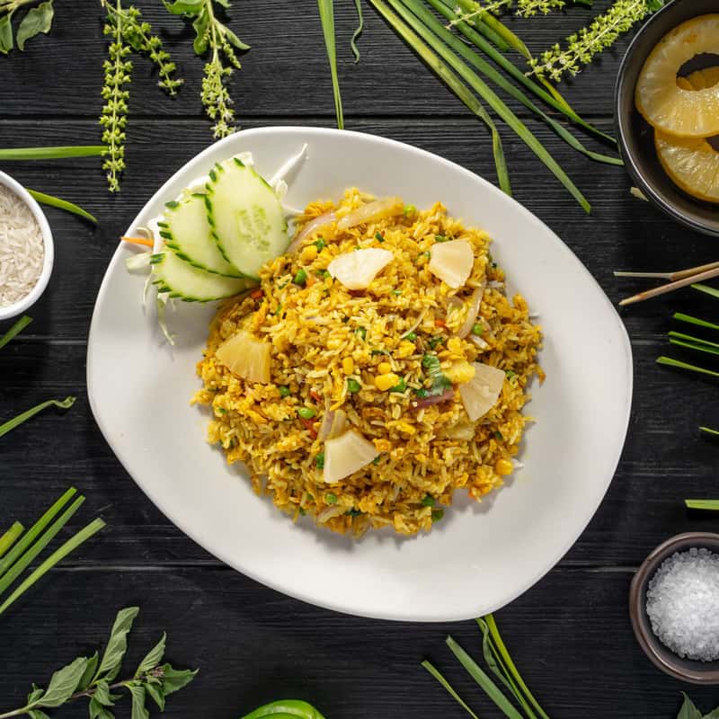 Stir-fried rice with eggs, pineapple, peas, onion, cashew nuts and curry powder on a white plate, on a black table decorate with vegetables around it.