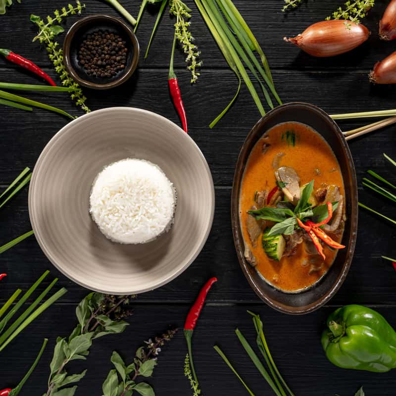Red curry paste with coconut milk, zucchini or eggplant, sweet pepper and sweet basil with a portion of rice on a black table decorate with vegetables around it.