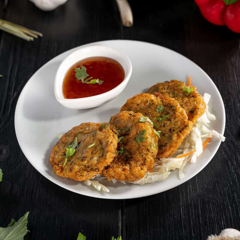 Three pieces of Thai Fish Cakes on a white plate with sweet and sour sauce on the side, all decorate with vegetables around the plate.