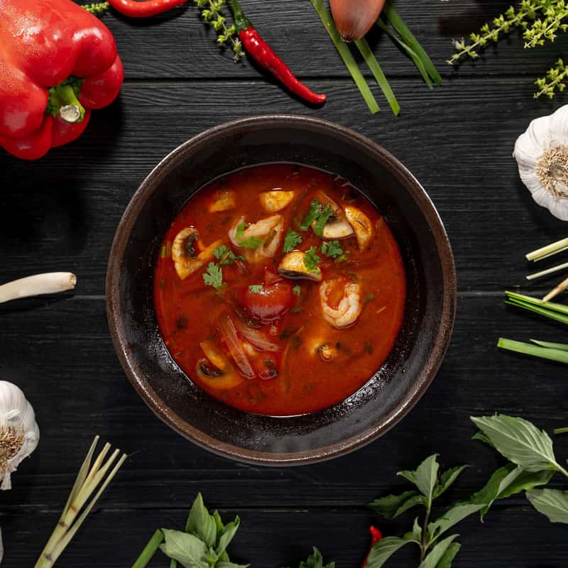 One portion of Tom Yam Soup on a black table decorate with vegetables around it.
