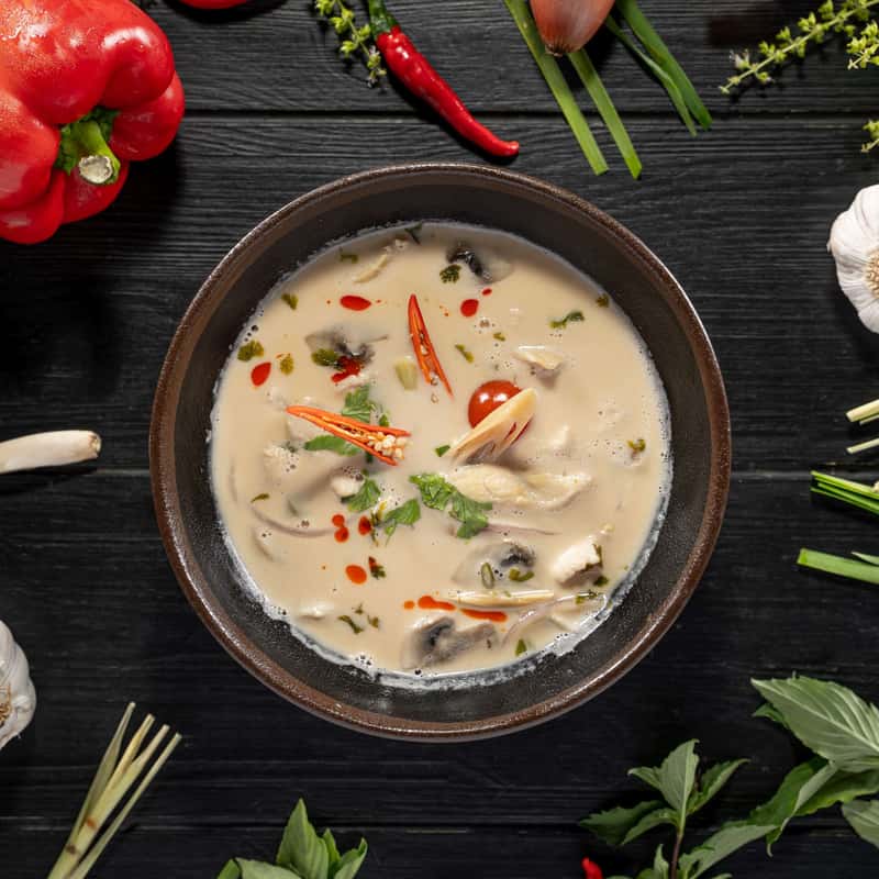 One portion of Tom Kha soup on a black table decorate with vegetables around it.