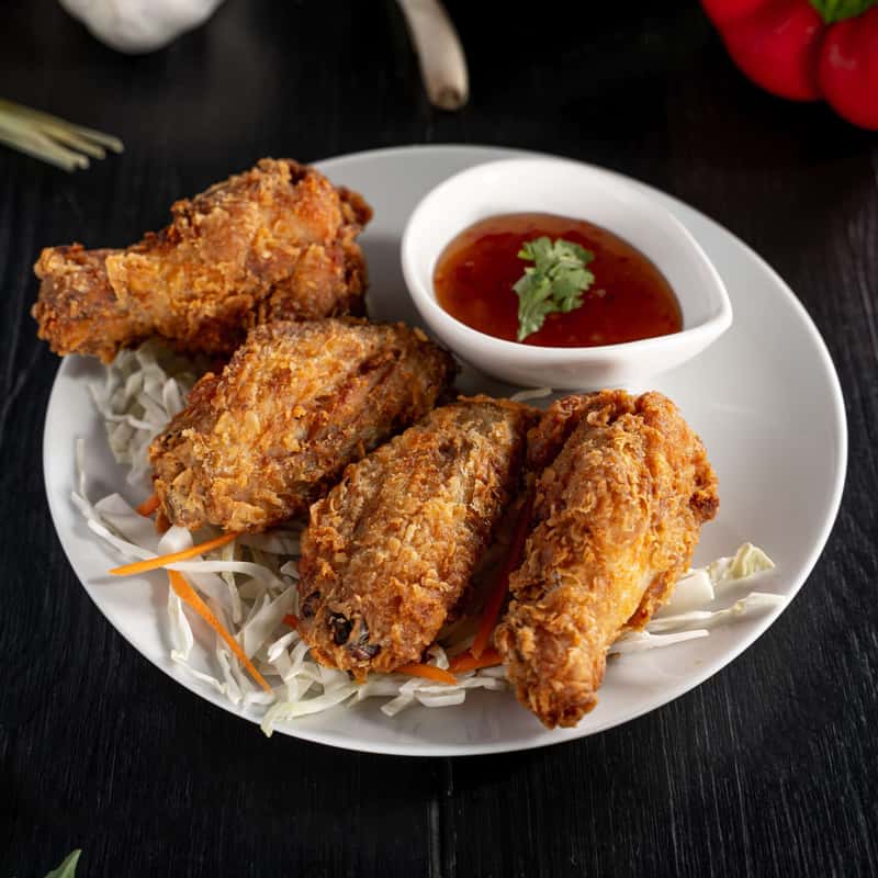 Four pieces of Chicken Wings on a white plate with sweet and sour sauce on the side, all decorate with vegetables around the plate.