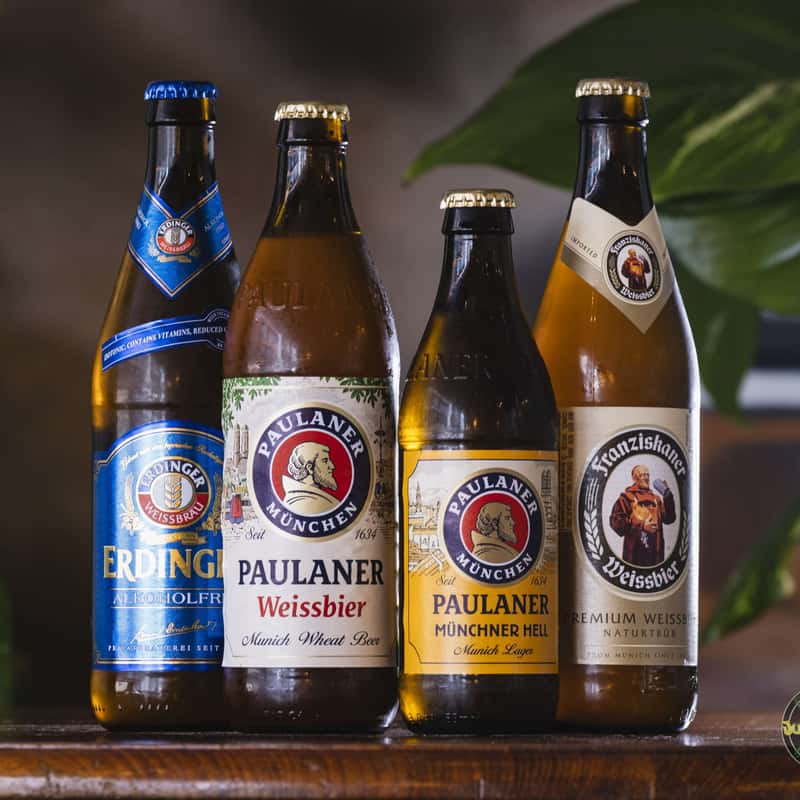 Four bottles of beer - Erdinger Non-Alcoholic, Paulaner Weissbier, Munchen, and Weissbier Franziskaner - arranged on a wooden table with a blurred dark background and a blurred plant on the left.