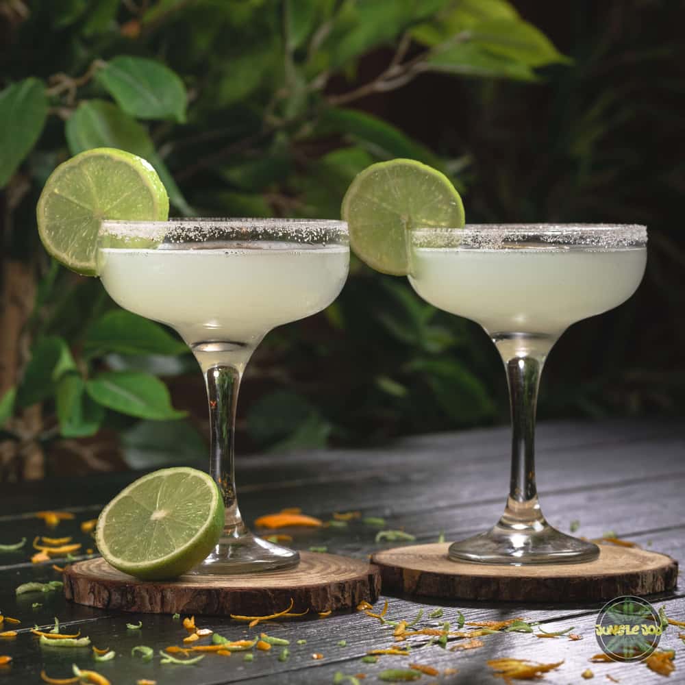 Two margarita cocktails are shown on a wooden table with a green tropical background. The drink is served in a manhattan glass with a salt rim and has a bright yellow-green color. The liquid is slightly cloudy and a slice of lime on the rim. The lighting is natural and creates a bright and vibrant atmosphere.