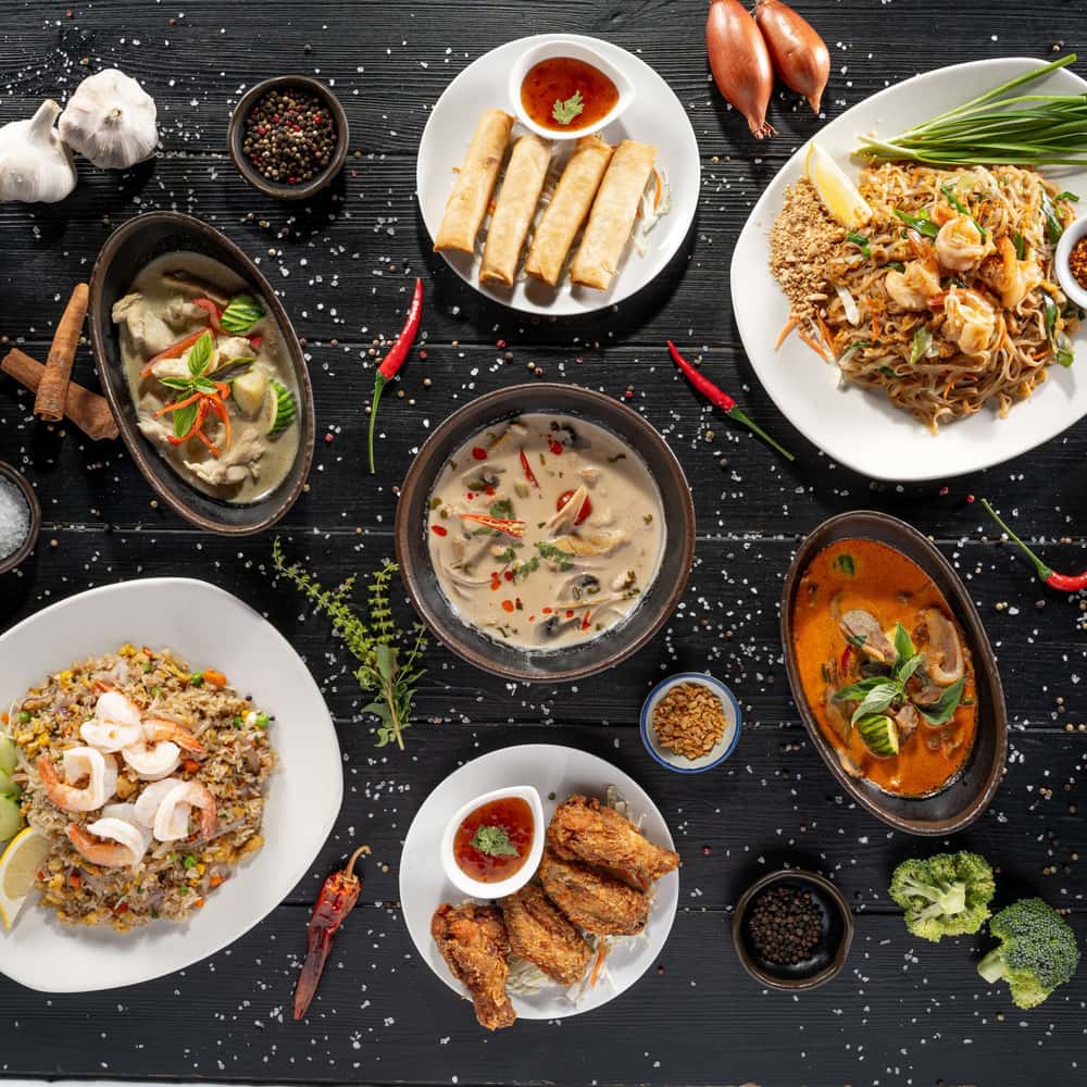 A photograph of a variety of Thai dishes presented on a black wooden table. In the center is a bowl of Thai soup, surrounded by spring rolls at the top, chicken wings at the bottom, red curry on the right, green curry on the left, pad Thai with prawns in the top right corner, and fried rice with prawns in the bottom left. The table is adorned with peppers, salt, broccoli, onion, and garlic. The image is taken from a top-down perspective, providing a clear view of the dishes' arrangement.