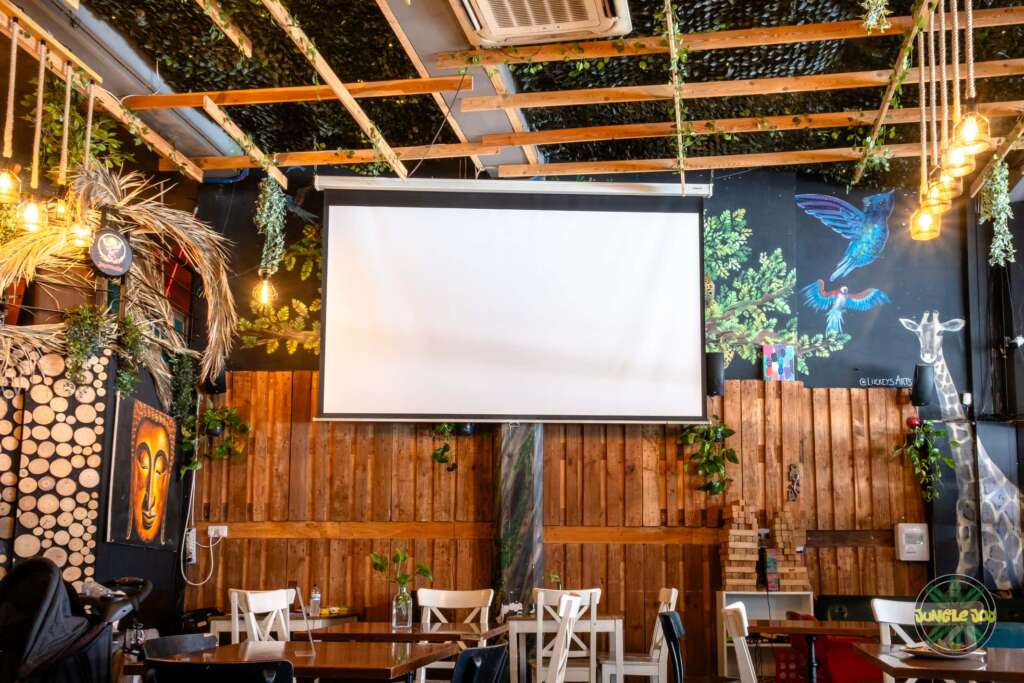 Jungle Joy Bar's vibrant inside room with wooden tables, chairs, and a large white screen for video projection. The space features yellow hanging lights, artistic wood panels, captivating mural art, and a green roof adorned with wooden tables and lush plants.