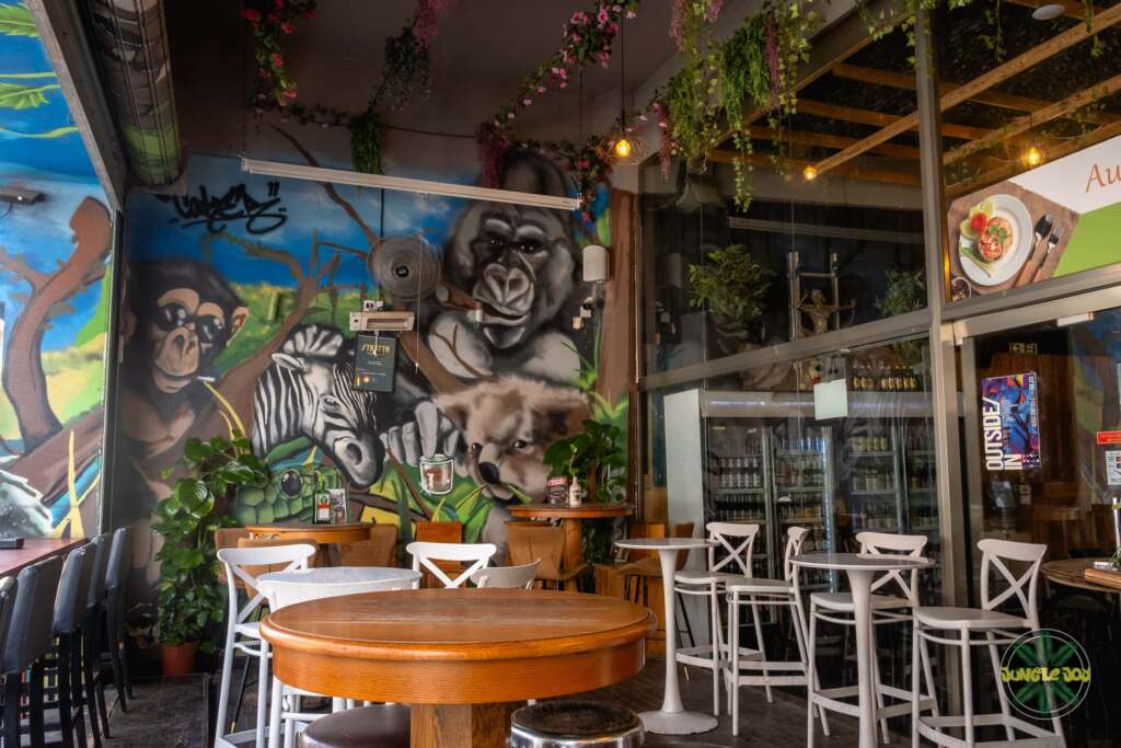Jungle Joy's inviting outdoor area with round wooden tables and chairs, adorned with captivating artistic mural art showcasing lively jungle animals. A perfect blend of nature and creativity, offering an immersive and unforgettable dining experience.