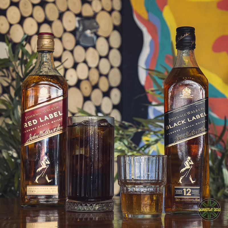 Impeccably Arranged Whiskey Delights on Wooden Table at Jungle Joy, Featuring Red Label, Whiskey Coke, and Black Label Double Shot, with Striking Murals in the Background.
