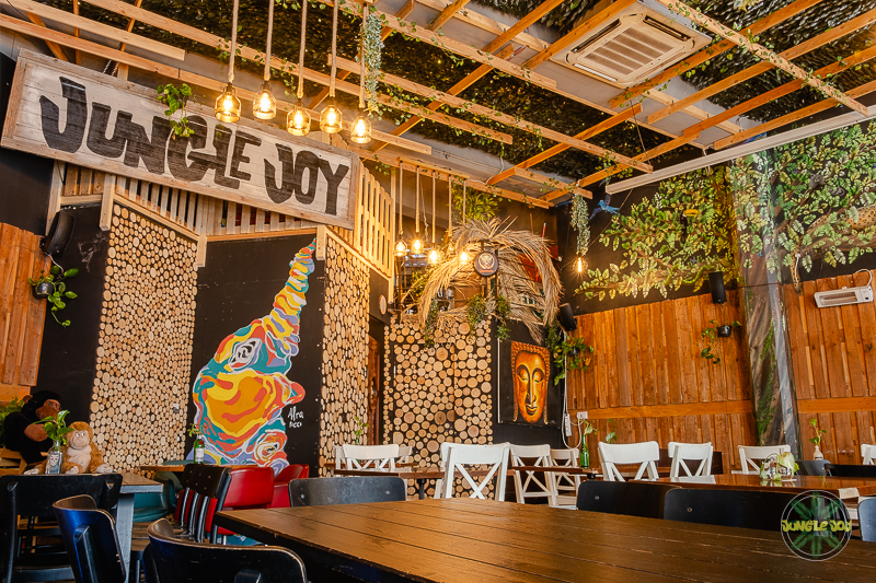 Spacious room with warm yellow ambient lighting, featuring wooden chairs and tables adorned with a decorative sign displaying 'Jungle Joy'. Hanging plants add a touch of greenery and natural charm to the space.