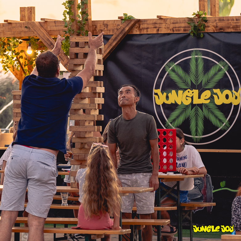A man carefully places a block atop the Giant Jenga tower as his friend observes from above. Nearby, a young girl sits beside the first man. In the background, a Jungle Joy Banner adds to the festive atmosphere