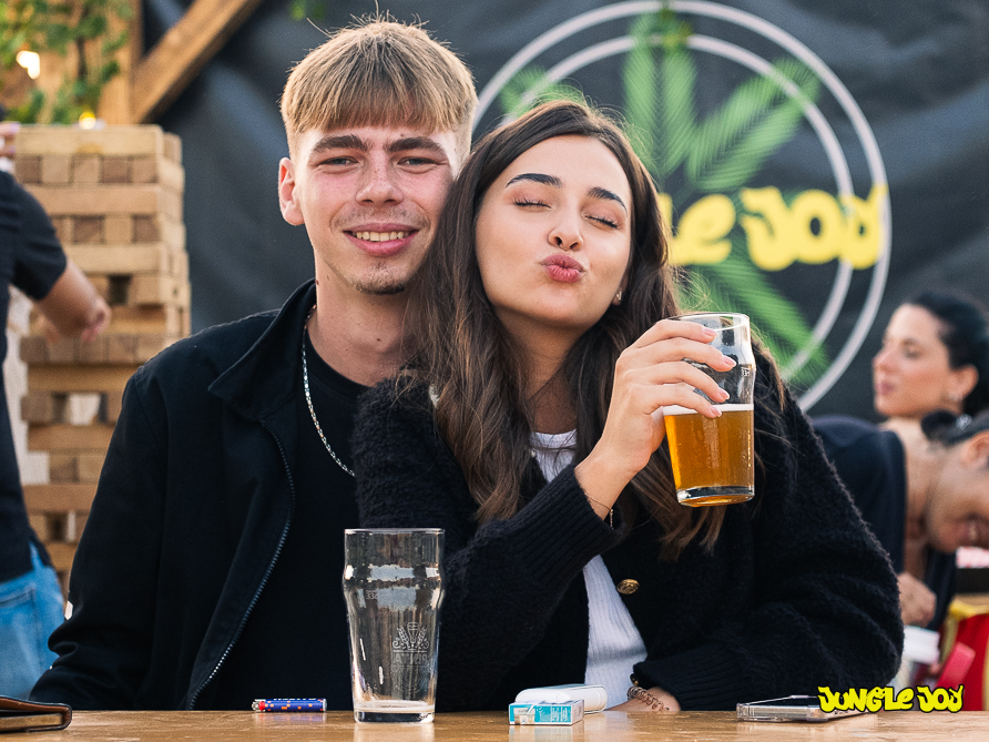 Young couple sits closely together, facing the camera. The guy, on the left, smiles while the girl blows a kiss, holding a beer glass with her left arm